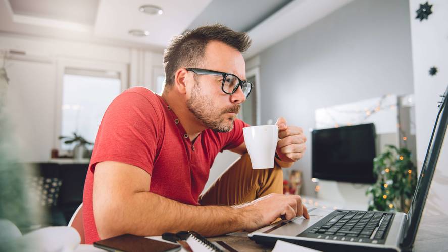 man in a shirt looking at a computer with a coffee in his hand - Adobe Stock