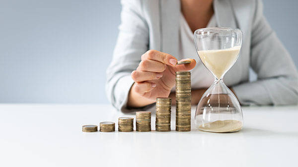 woman in suit adding coins on a pile next to an hourglass - Andrey Popov-AdobeStock