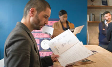 Un homme vêtu d'un costume regarde attentivement un document dans un espace de bureau, tandis que d'autres personnes en arrière-plan examinent également des papiers.