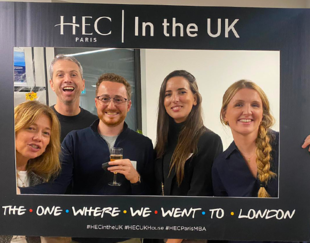 Five smiling people posing with a photo frame that reads HEC Paris In the UK - The One Where We Went to London' at an event in London