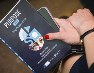woman's hands holding a program for an event titled "Purpose Day". The program's cover is dark gray featuring a colorful mask logo and states "PURPOSE DAY 4: Bring purpose to life" dated March 8, 2024
