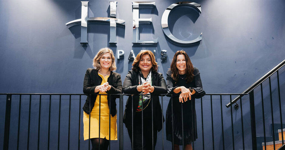 Three smiling women standing in front of a large HBC logo