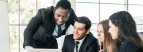 Des hommes d'affaires africains se tenant à l'arrière de collègues caucasiens souriants regardant sur leur ordinateur portable lors d'une réunion d'un groupe d'affaires interne avec un tableau blanc vide. Diversité de la coopération des hommes d'affaires.