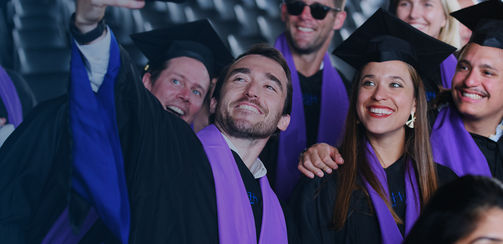 Un groupe de diplômés prenant un selfie, souriant et ayant l'air heureux. Ils portent des toges académiques noires et des chapeaux avec des écharpes violettes. L'arrière-plan montre une zone de sièges, indiquant une cérémonie de remise des diplômes à l'intérieur